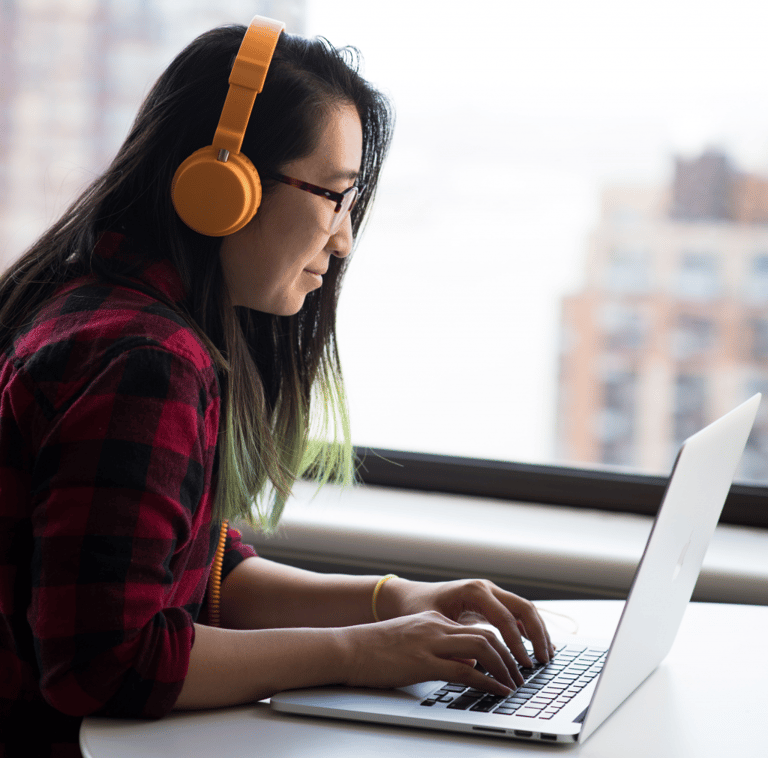 Image source: Christina @Wocintechchat.com via Unsplash, photo of smiling woman wearing yellow headphones while working at a laptop computer near a large window.