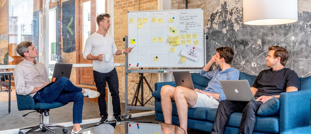 Group of young male business associates gather around a whiteboard. Image by Austin Distil via Unsplash.